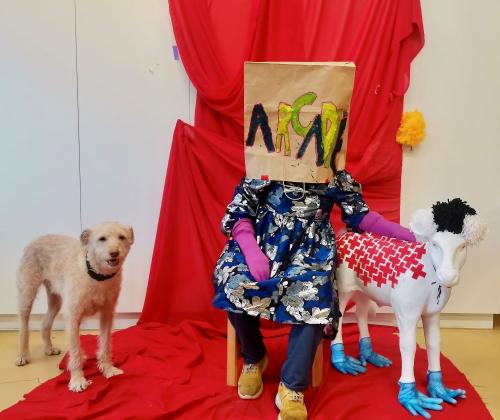 kunstenares zit met papieren tas over het hoofd op een stoel voor een rood doek, links een hond, rechts een even grote replica van een schaap 